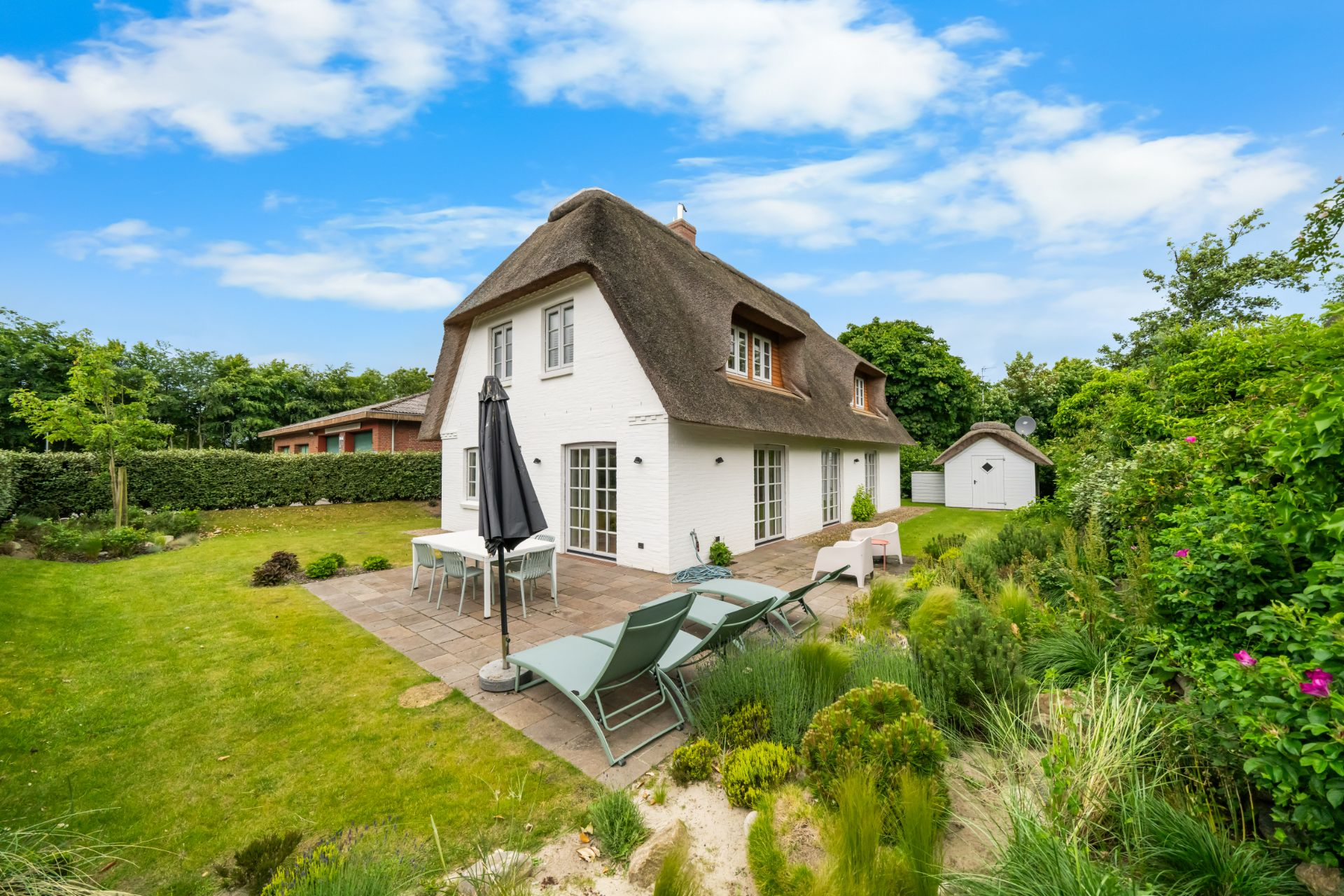 Weißes Haus mit Reetdach, Terrasse, Gartenmöbeln und grünem Garten bei blauem Himmel.