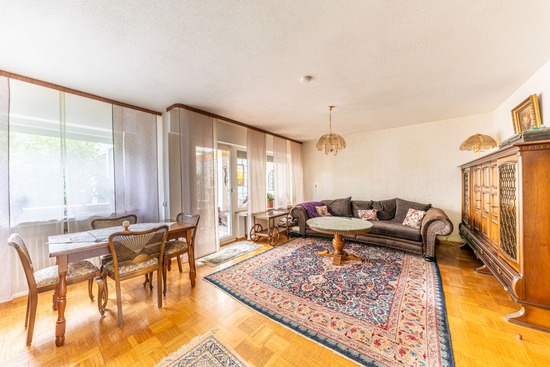 Helles Wohnzimmer mit Teppich, Sofa, Esstisch und großen Fenstern.