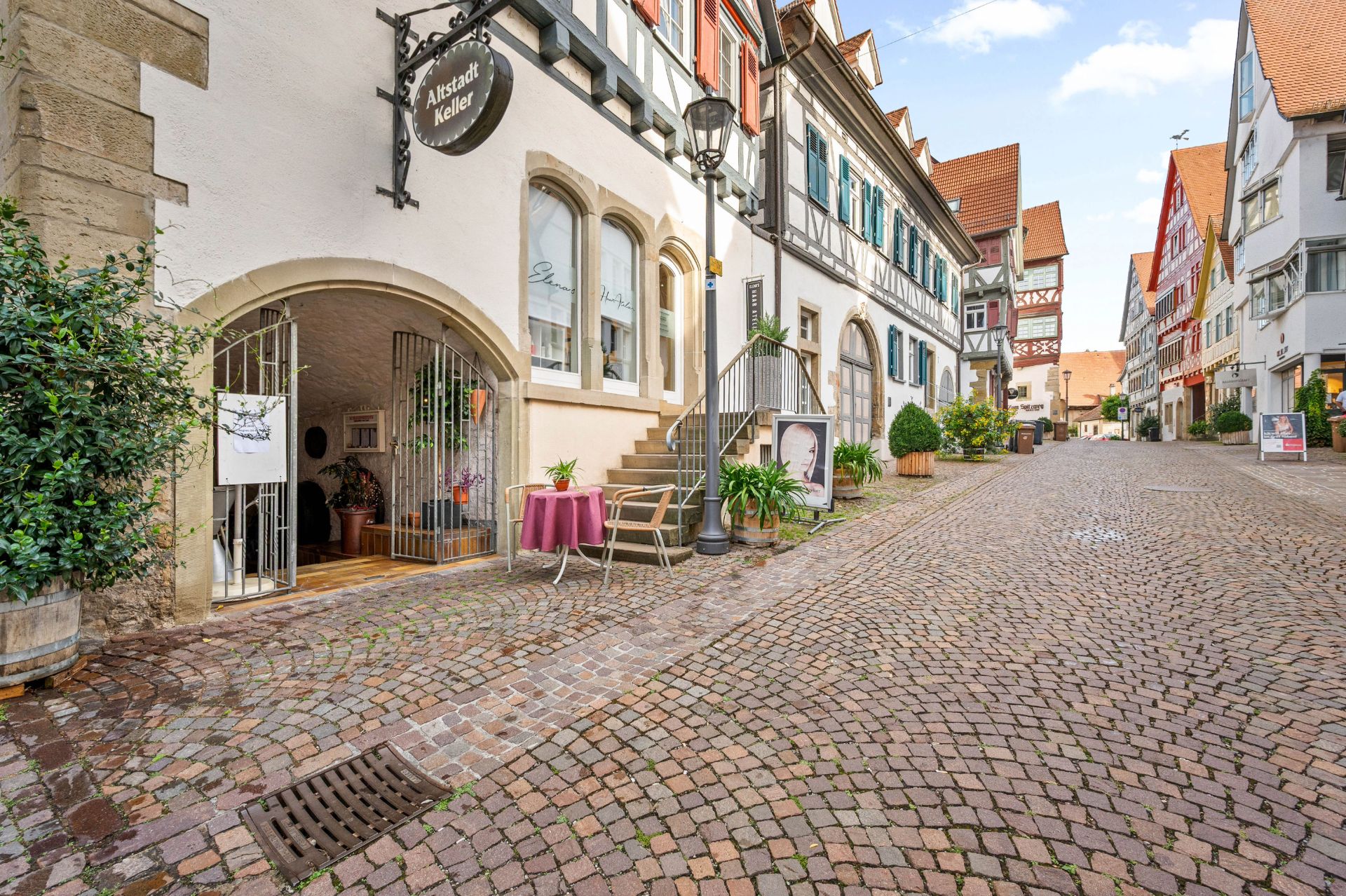 Gepflasterte Gasse mit Fachwerkhäusern, Pflanzen und einem kleinen Tisch mit zwei Stühlen vor einem Gebäude.