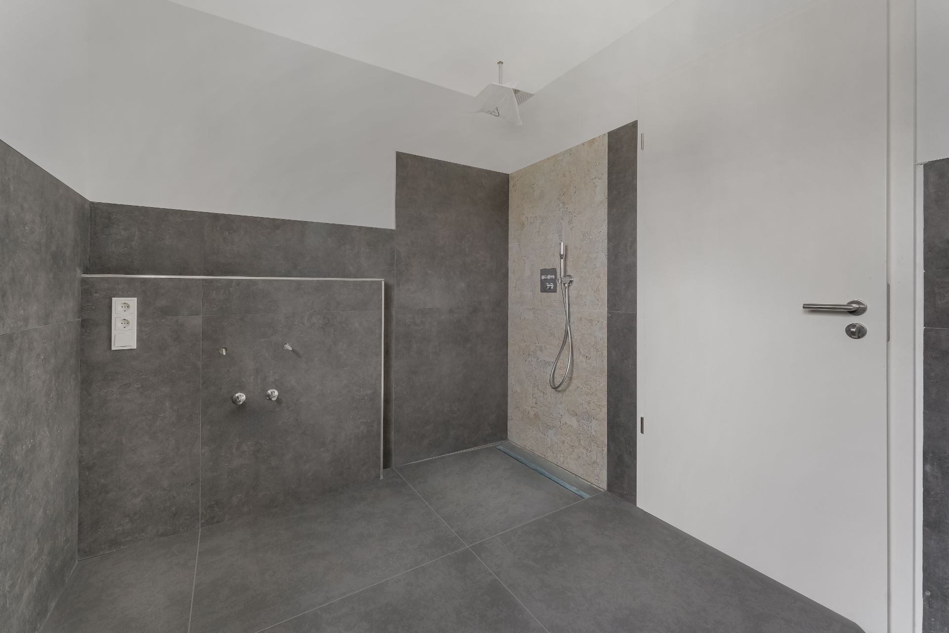 Modernes Badezimmer mit grauen Fliesen, Dusche, weißer Tür und Steckdosen an der Wand.