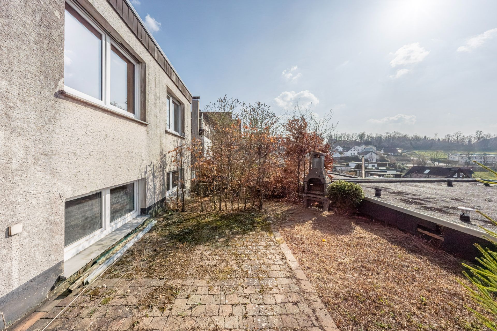 Kleiner Garten mit Terrasse neben einem Wohnhaus, einige Sträucher und ein gemauerter Grill sind sichtbar.