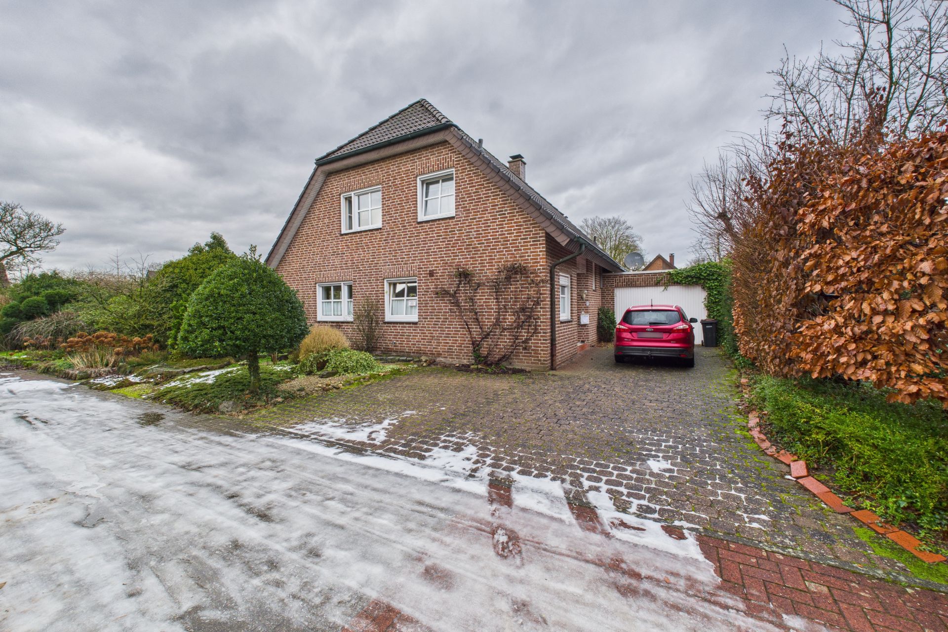Backsteinhaus mit Einfahrt, rotem Auto und winterlichem Vorgarten.