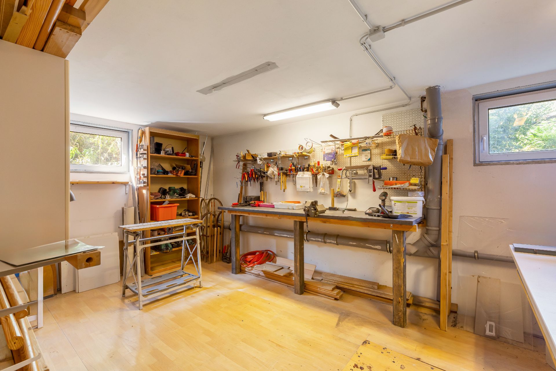 Helles Arbeitszimmer mit Werkbank, Regalen, Werkzeugen und zwei Fenstern.