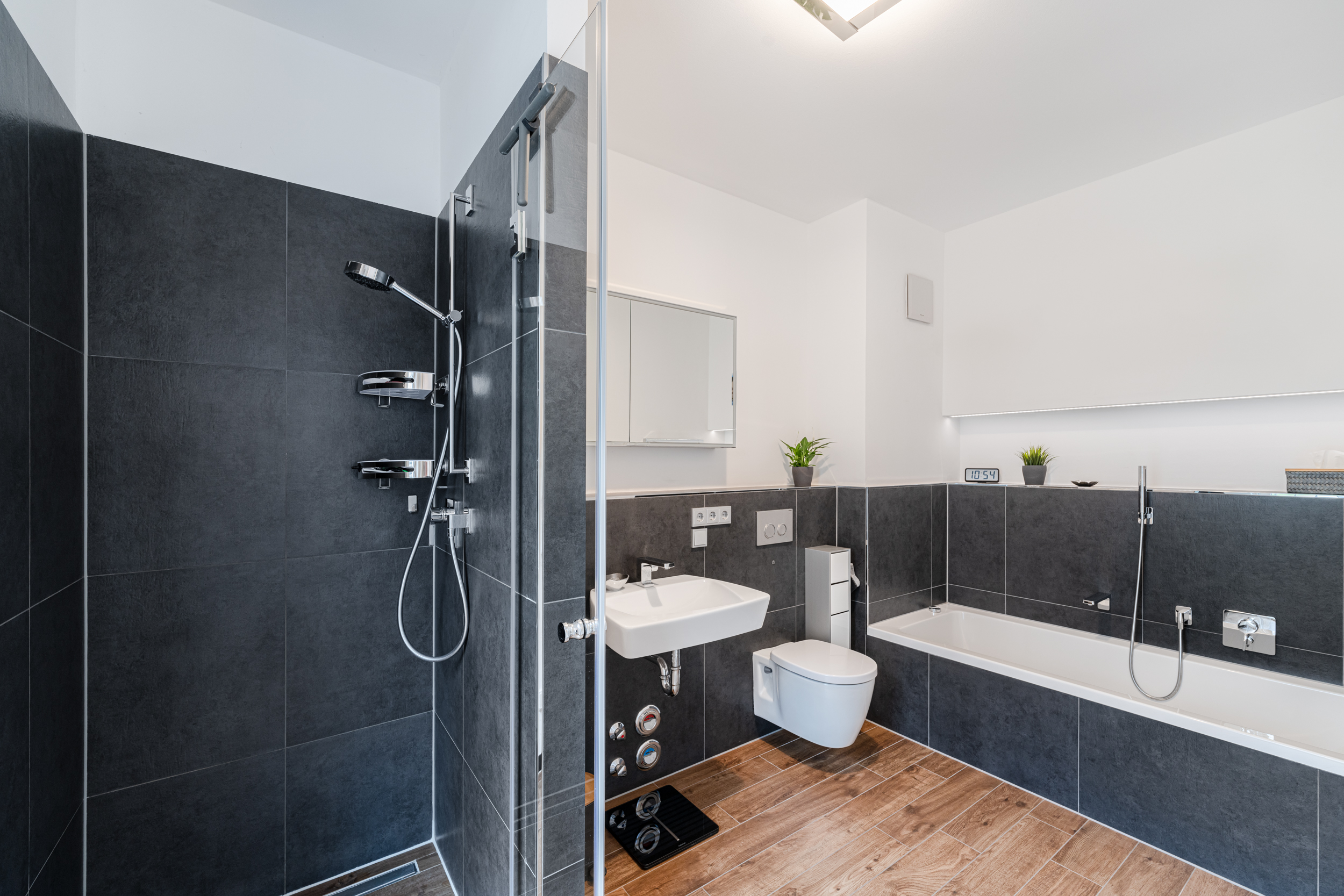 Modernes Badezimmer mit Dusche, Badewanne, Waschbecken und WC, dunkle Fliesen und Holzboden.