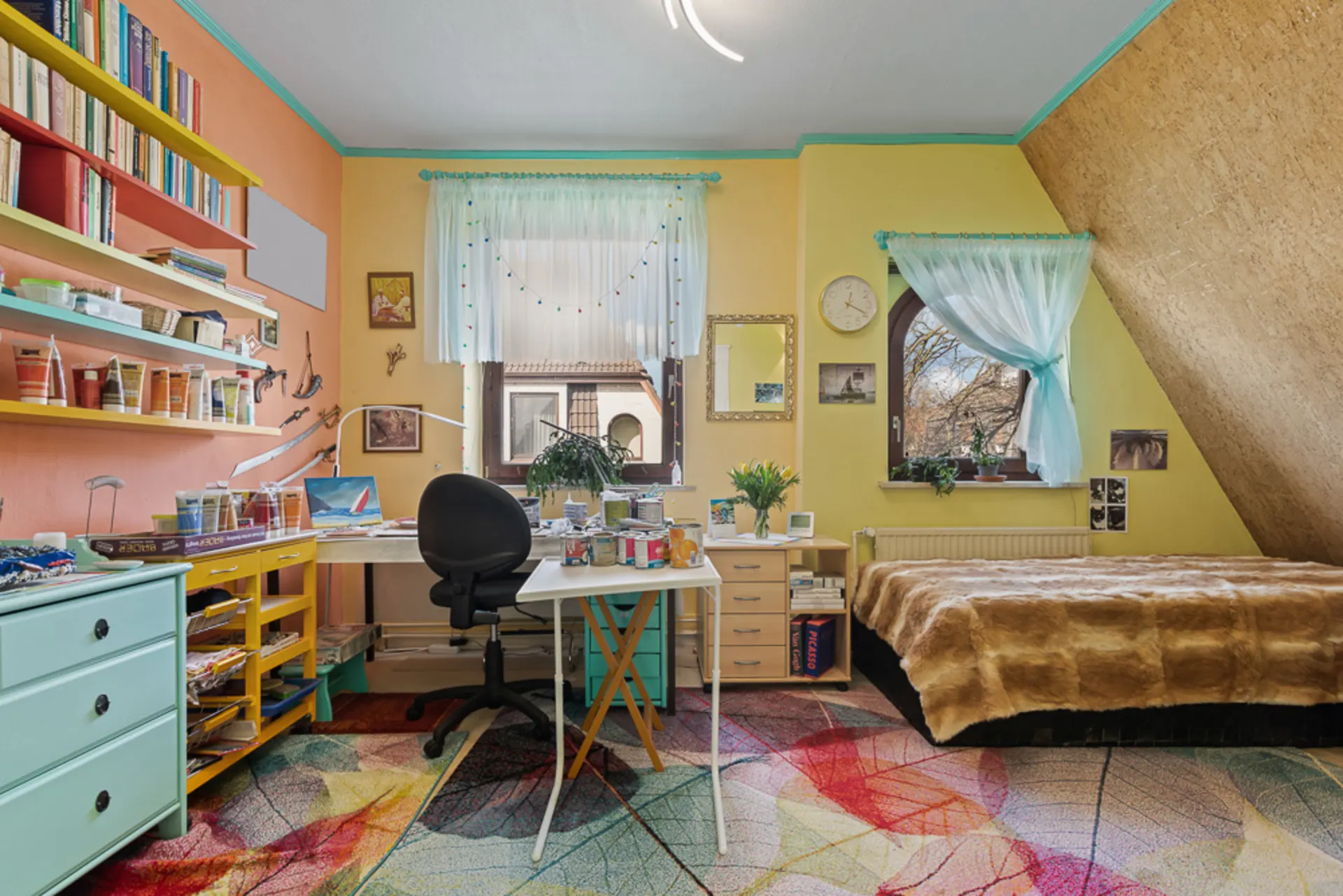 Wohnzimmer mit Schreibtisch, Bücherregalen, bunten Wänden, Bett und Teppich mit Blättermuster.
