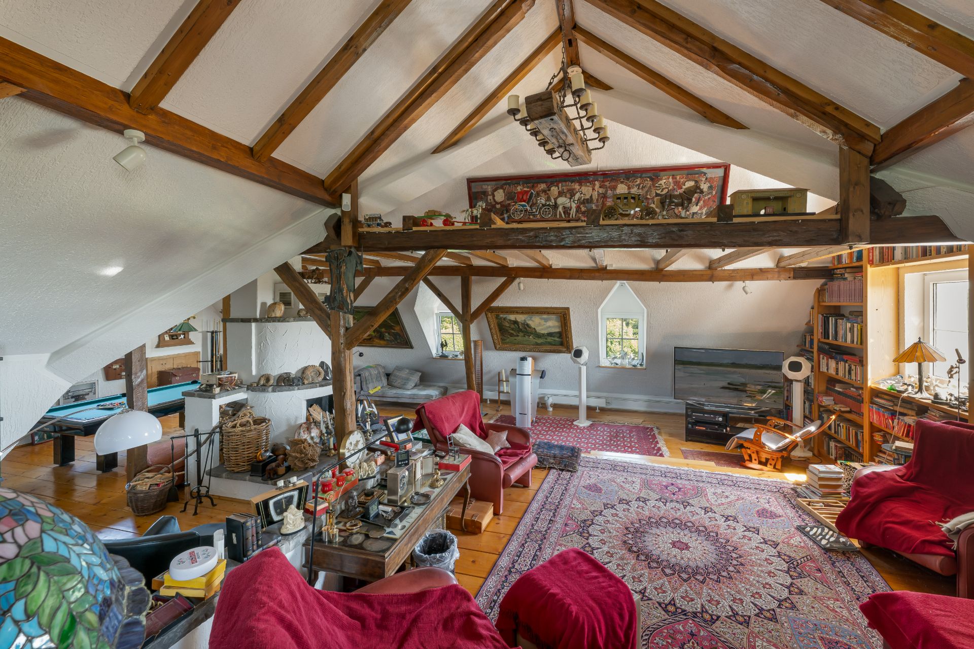 Wohnzimmer mit Holzbalkendecke, roten Sesseln, Teppich, Regal und Billardtisch.