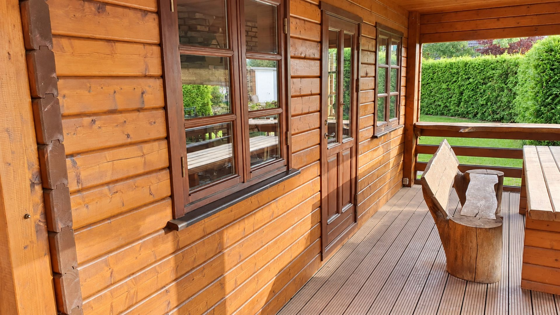 Überdachte Holzterrasse mit Tisch und Bank, daneben Fenster und Tür, im Hintergrund grüne Hecke.