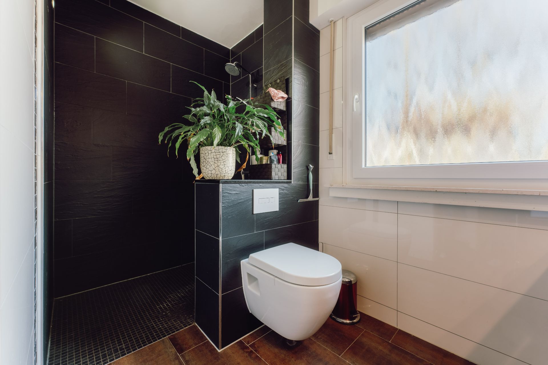 Modernes Badezimmer mit schwarzer Dusche, Hänge-WC, Pflanze und großem Fenster.