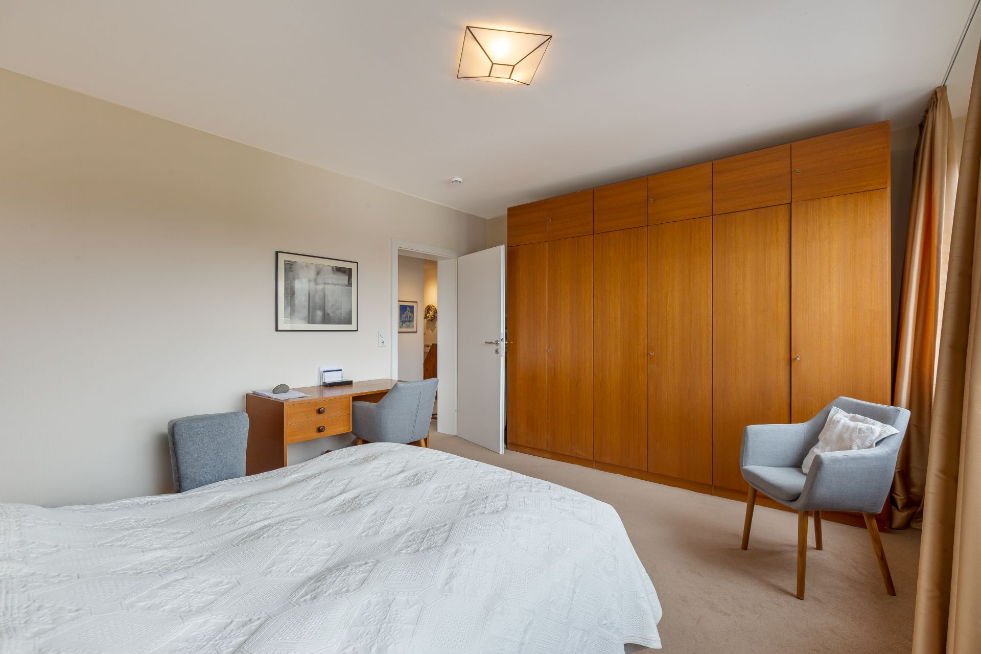 Helles Schlafzimmer mit großem Holzschrank, Bett, Schreibtisch und zwei grauen Sesseln.