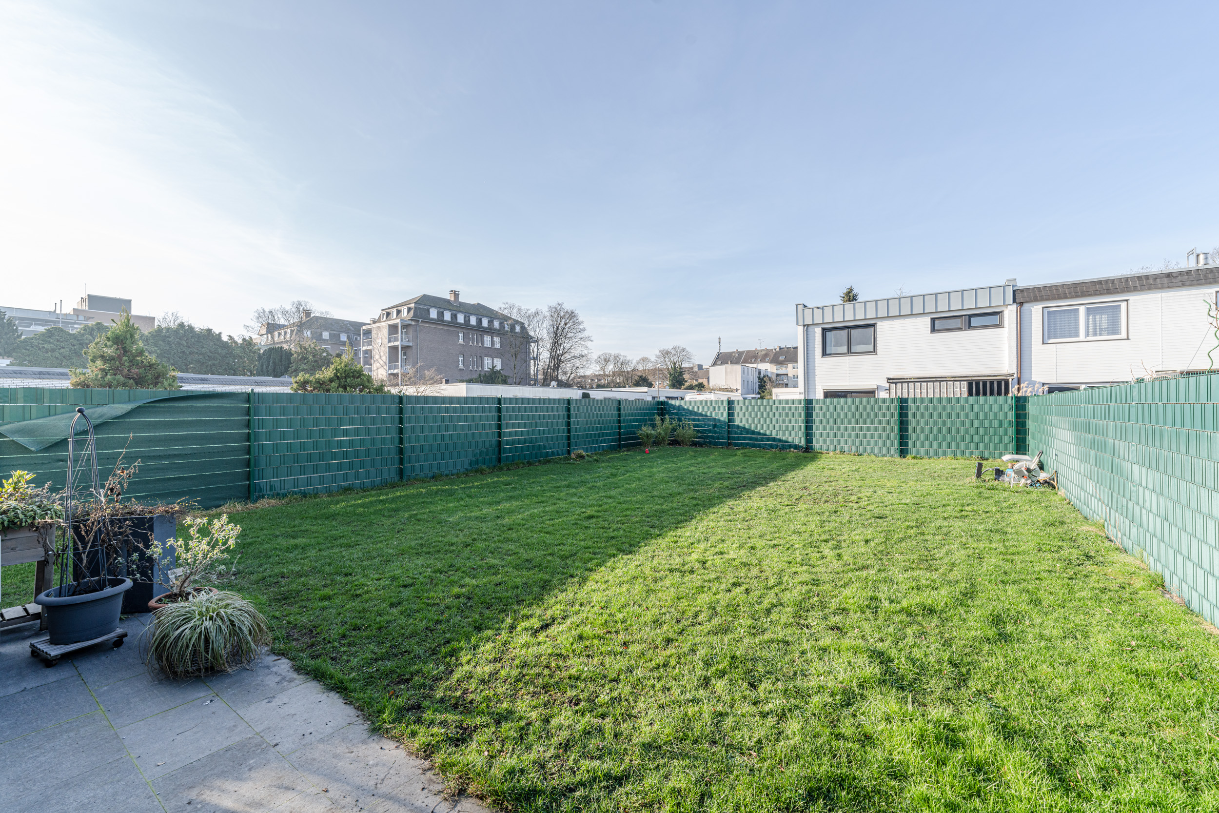 Garten mit grünem Rasen, wenigen Pflanzen und grünem Zaun, umgeben von Wohnhäusern.