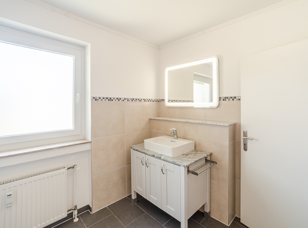 Modernes Badezimmer mit Fenster, Waschbecken, beleuchtetem Spiegel und hellen Fliesen.