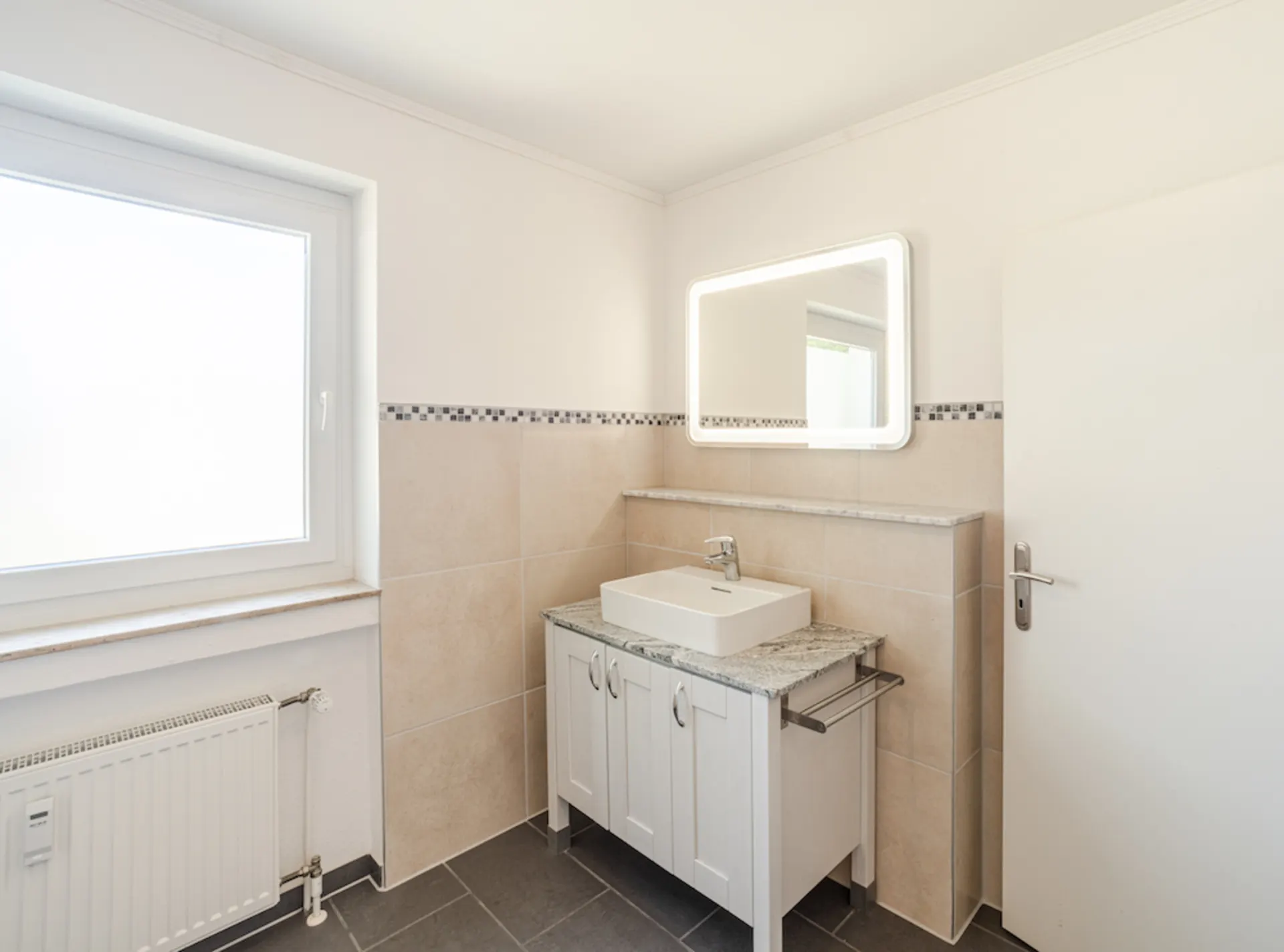 Modernes Badezimmer mit Fenster, Waschbecken, beleuchtetem Spiegel und hellen Fliesen.
