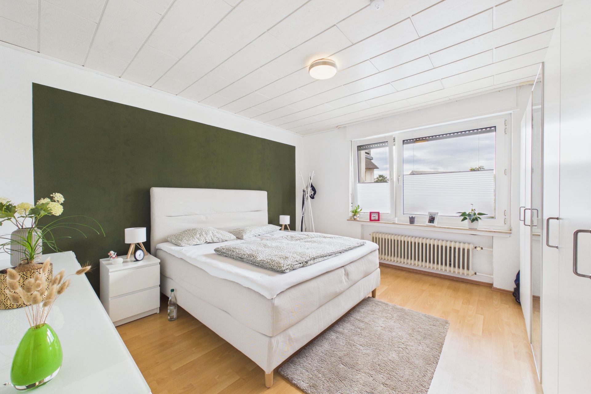 Helles Schlafzimmer mit großem Bett, grünem Akzentwand, Fenster und Holzfußboden.