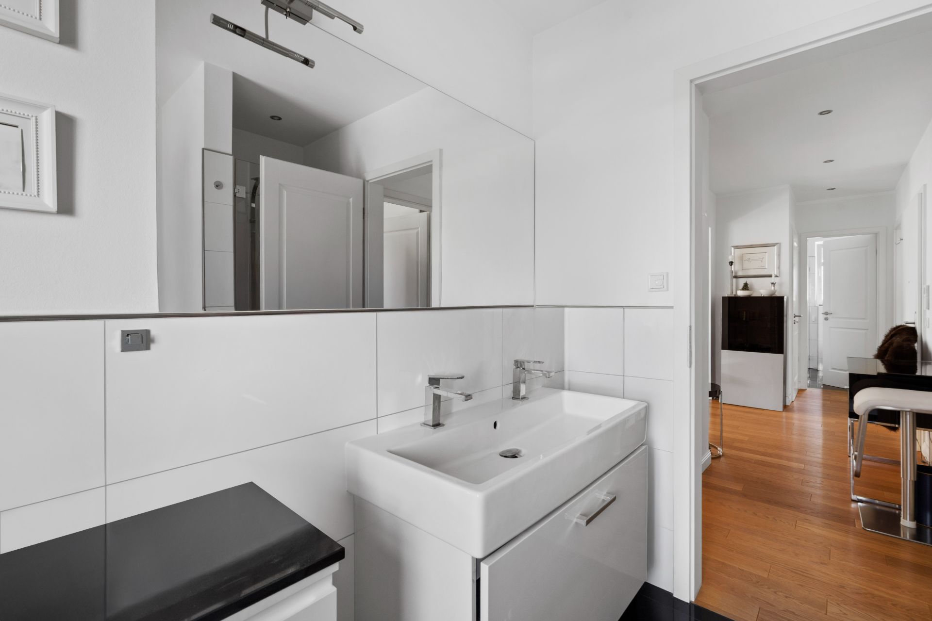 Modernes Badezimmer mit weißem Waschbecken, Spiegel und Blick in einen Flur mit Holzboden.