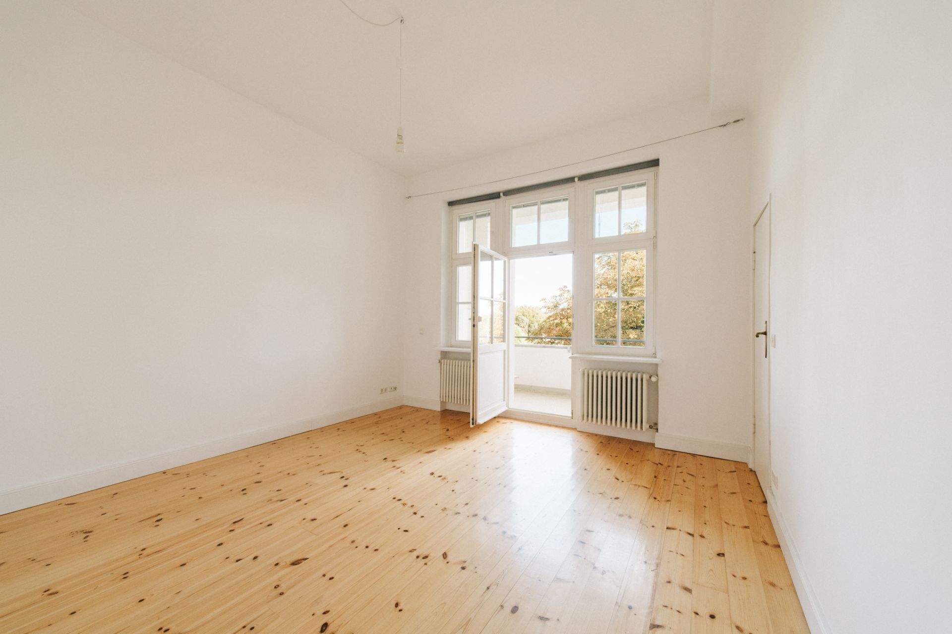 Leerer Raum mit Holzboden, weißen Wänden und großen Fenstern mit offener Tür zum Balkon.