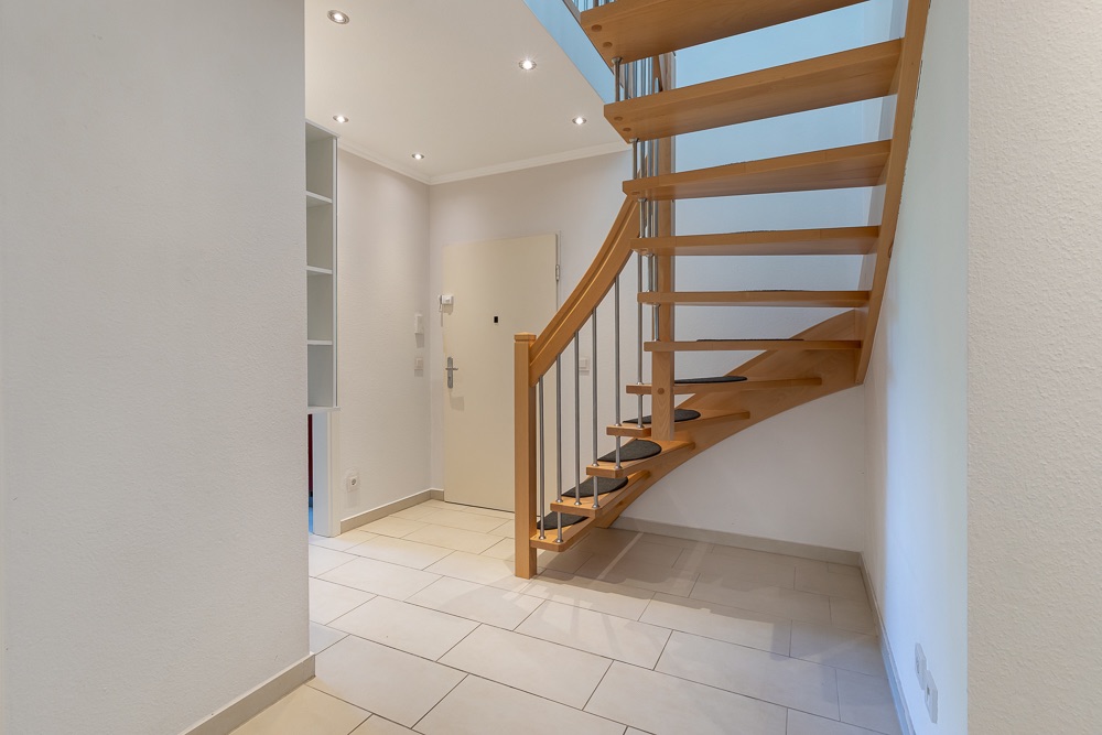 Heller Flur mit moderner Holztreppe, weißen Wänden und Fliesenboden.