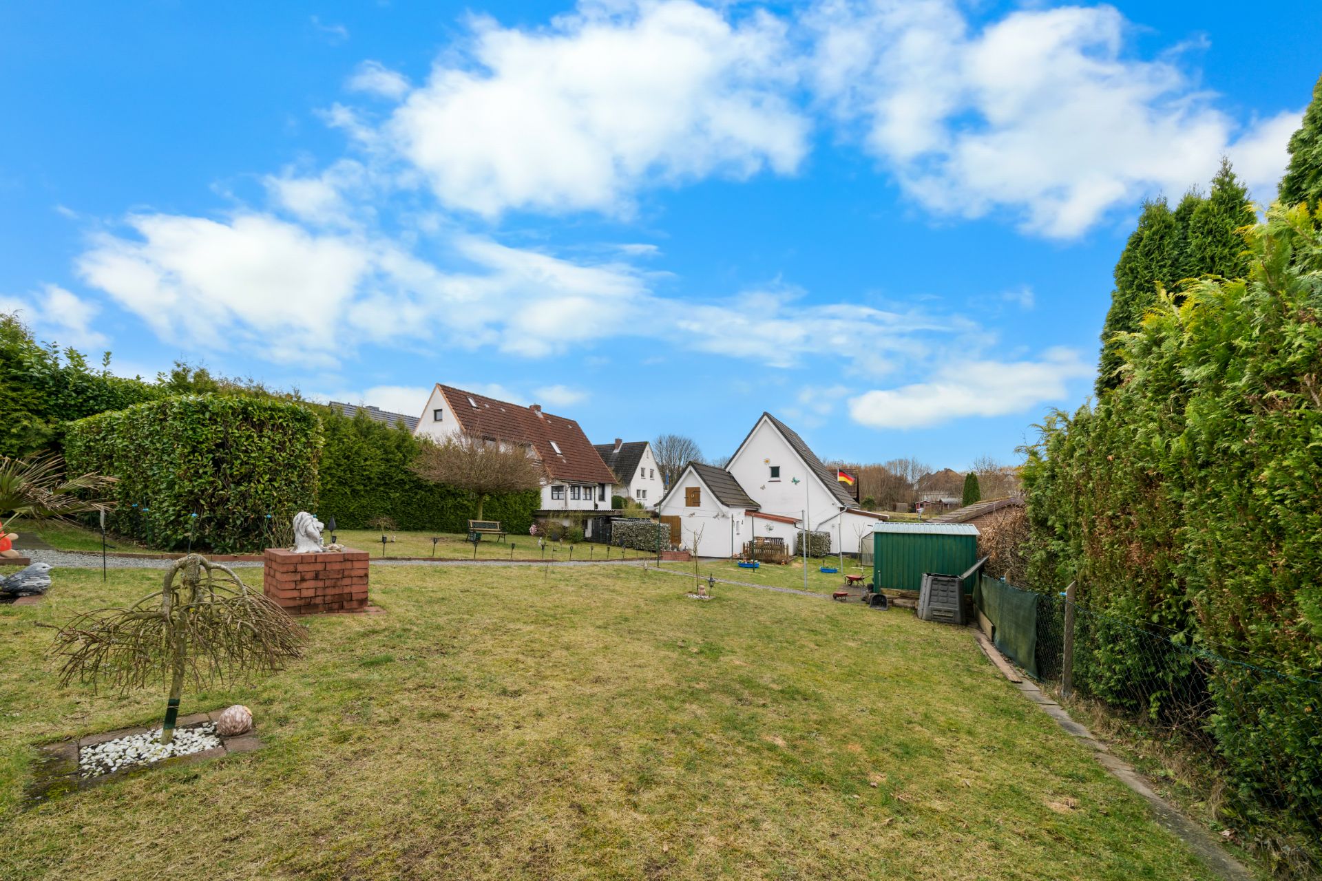 Großer Garten mit Rasen, einigen Sträuchern und Häusern im Hintergrund bei blauem Himmel.