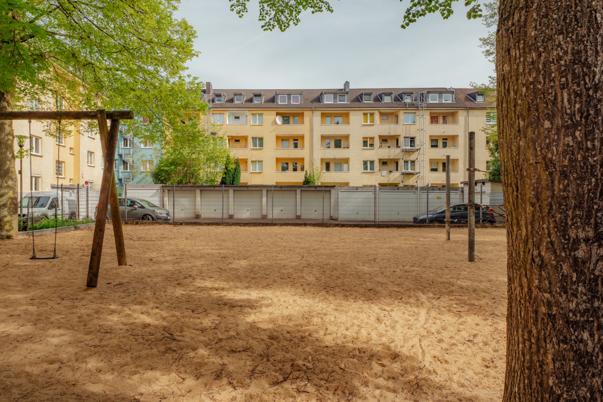 Sandiger Spielplatz mit Schaukel und Bäumen vor einem mehrstöckigen Wohnhaus.