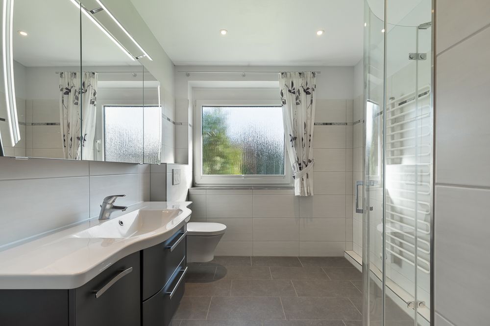 Modernes Badezimmer mit großem Spiegel, Waschbecken, Toilette, Fenster und Glasdusche.