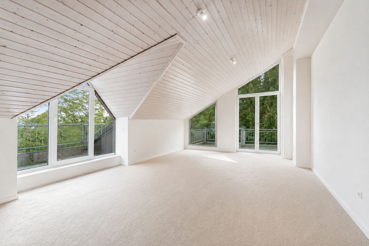 Heller Dachgeschossraum mit schräger Holzdecke, großen Fenstern und Blick ins Grüne.