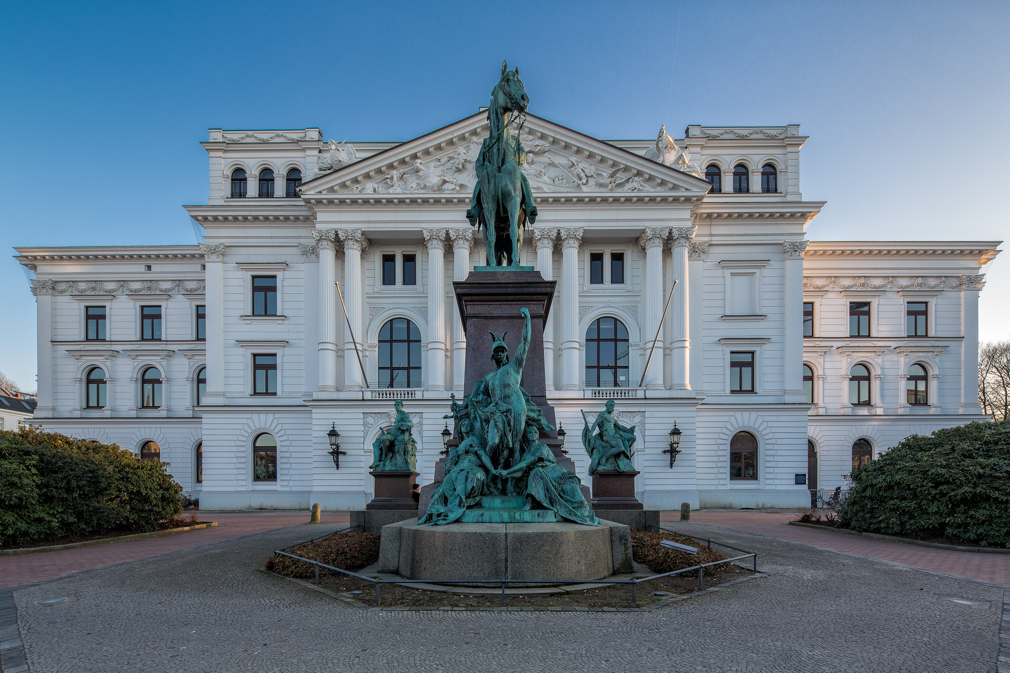 Vor einem weißen, prunkvollen Gebäude steht ein großes Reiterstandbild mit mehreren Figuren.