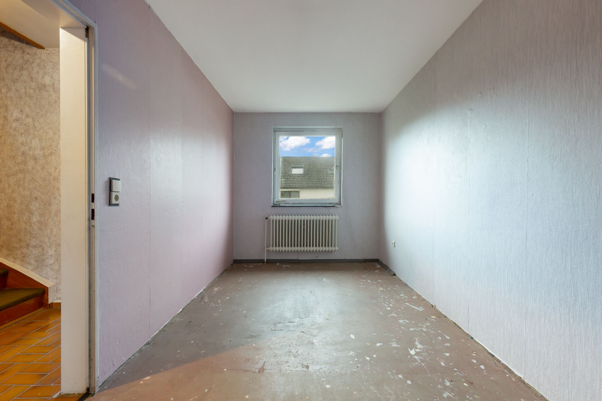 Leerer Raum mit einem Fenster, Heizkörper und abgenutztem Boden, links eine offene Tür zum Flur.