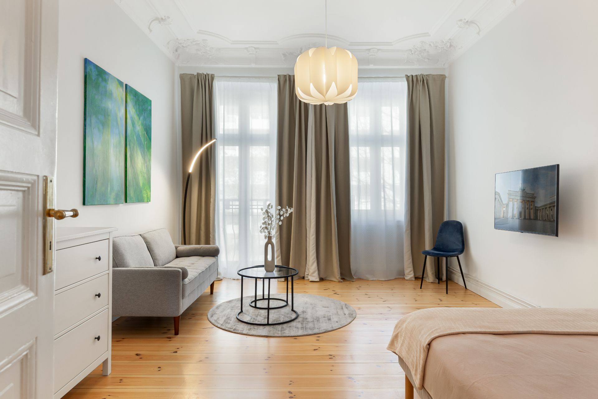 Helles Wohnzimmer mit Sofa, Teppich, Tisch, Bett, Vorhängen und Wandbild.