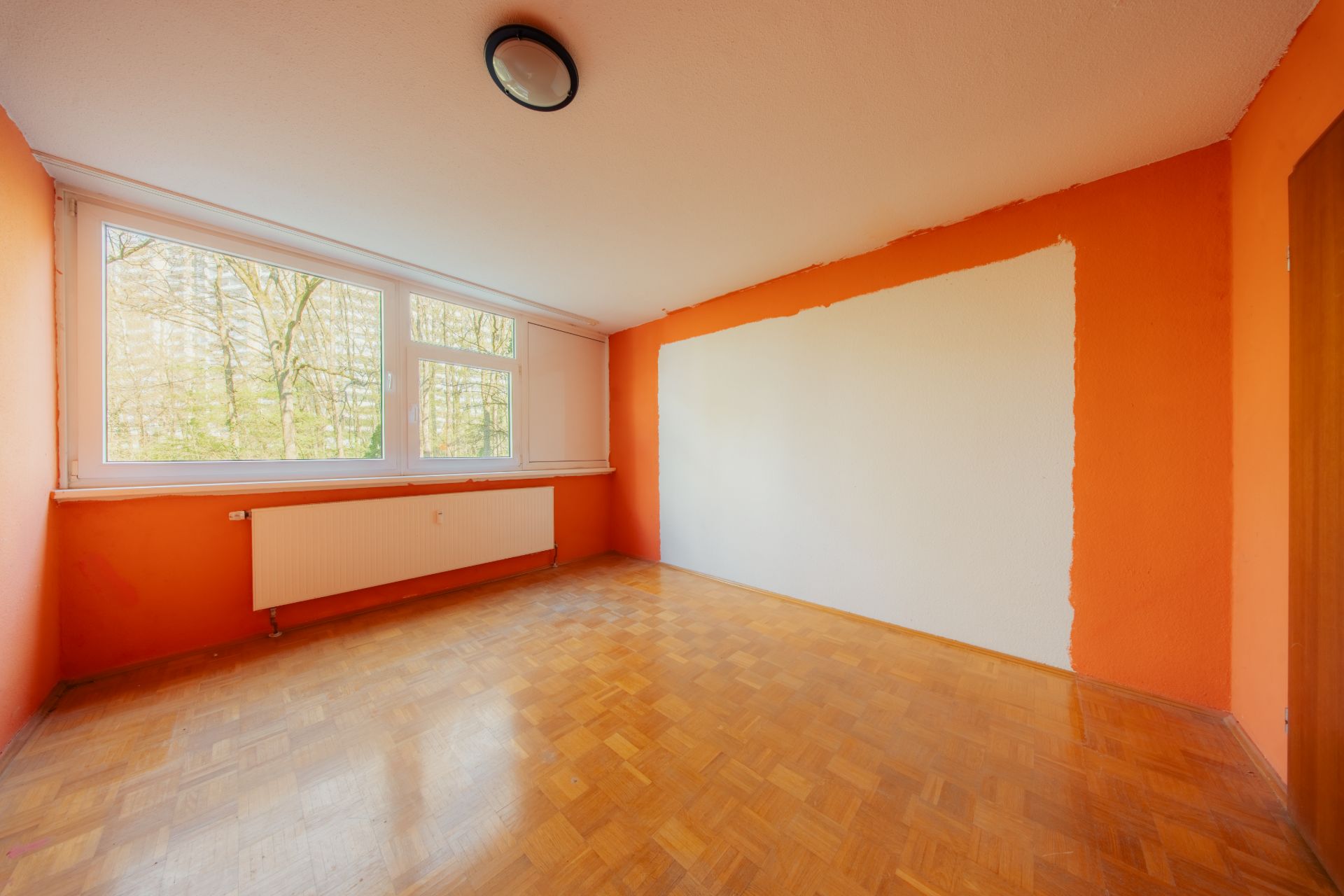 Leeres Zimmer mit Parkettboden, orangenen Wänden und großem Fenster mit Blick ins Grüne.