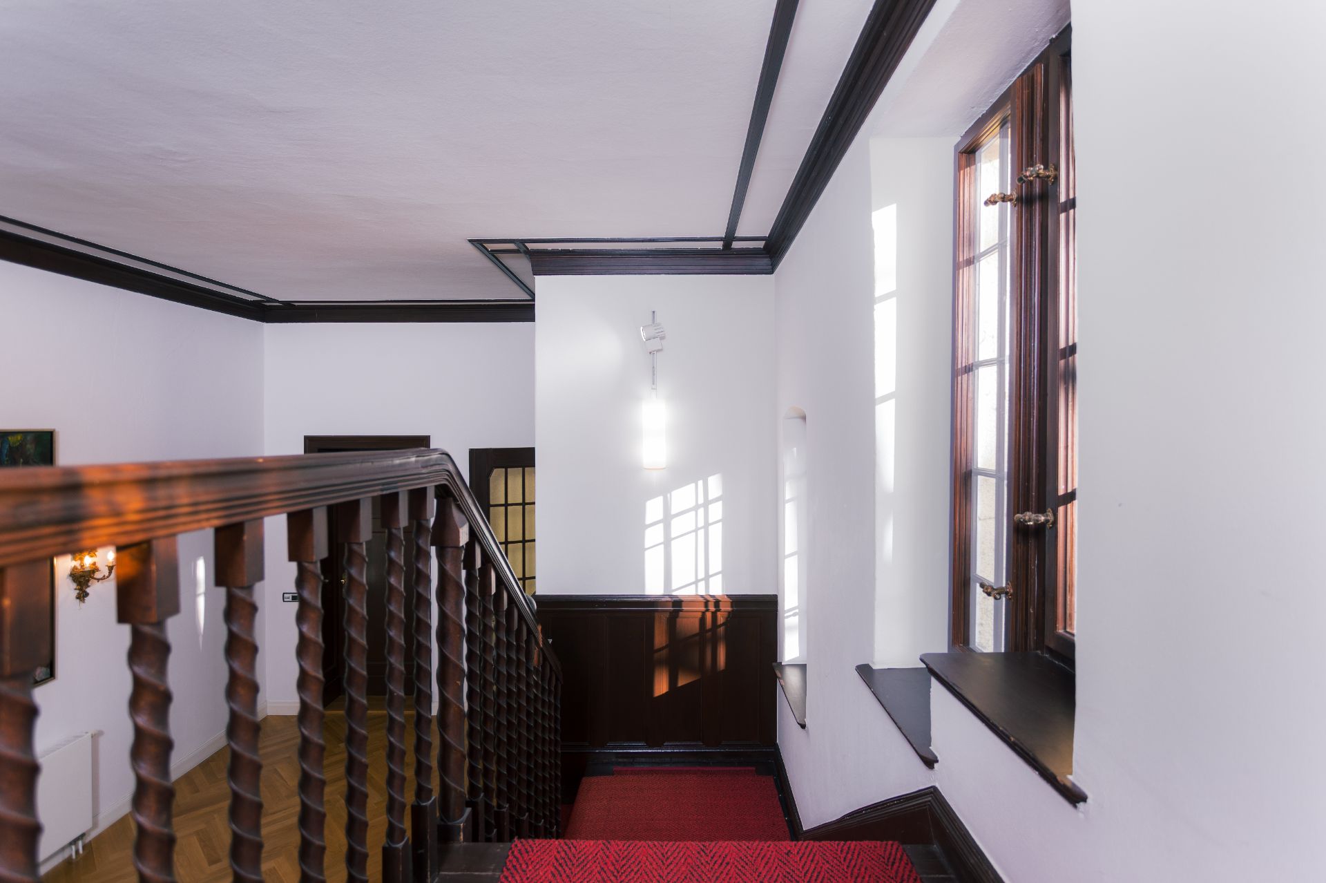 Treppe mit Holzgeländer und rotem Teppich, große Fenster an der rechten Wand.