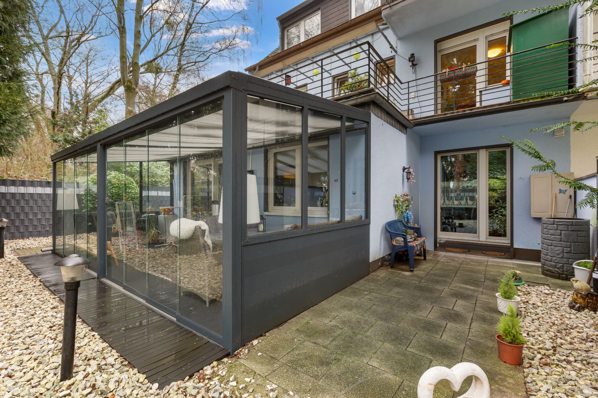 Wintergarten mit Glaswänden auf Steinterrasse neben einem blauen Wohnhaus mit Balkon.
