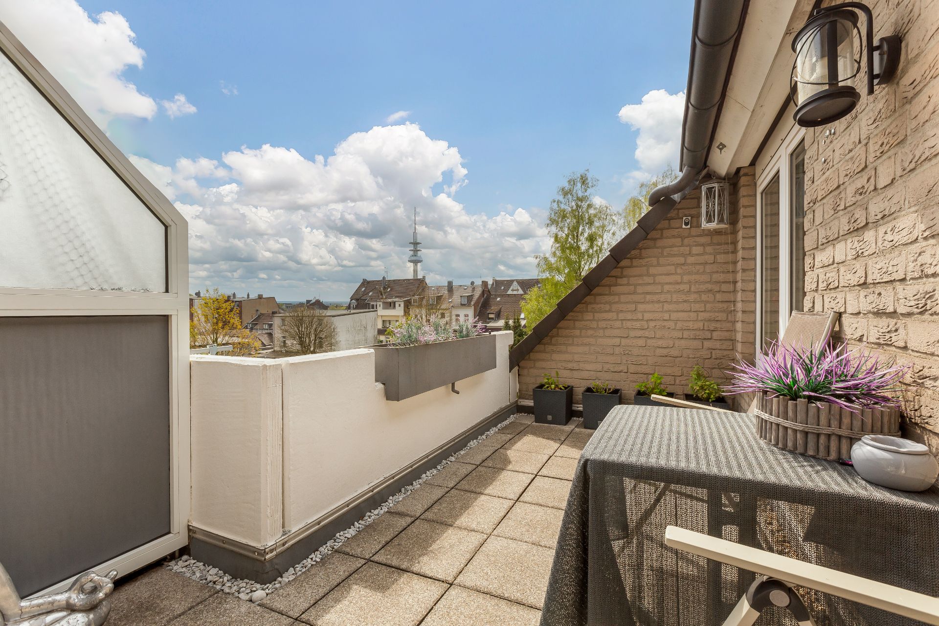 Helle Dachterrasse mit Pflanzen, Tisch, Stuhl und Blick auf Nachbarhäuser unter blauem Himmel.