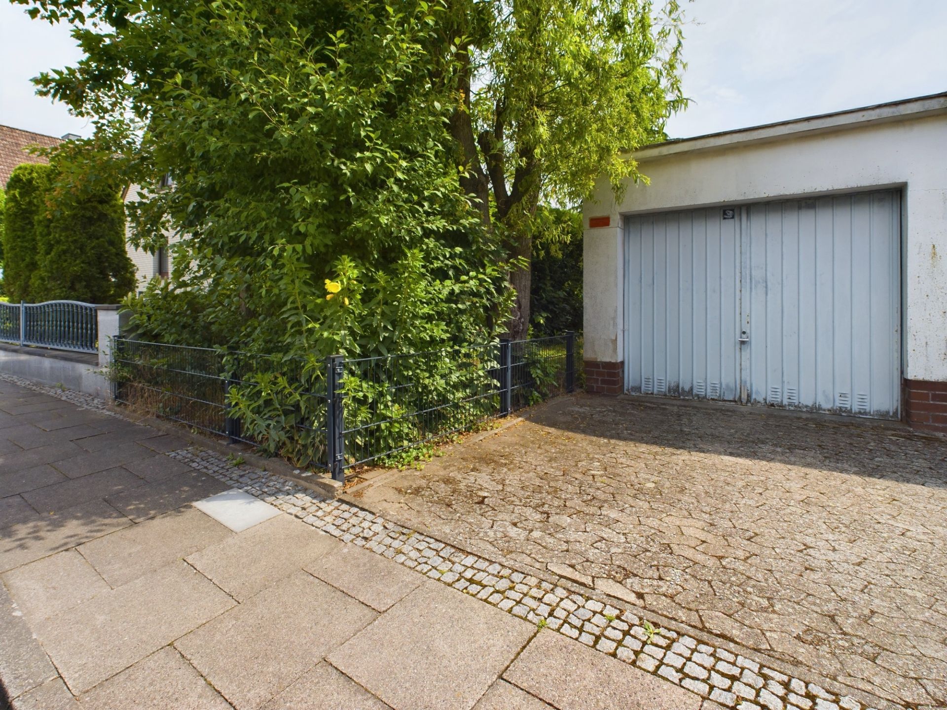 Garage mit verschlossenem Tor neben einem Garten mit Busch und Baum an einer Gehwegkante.