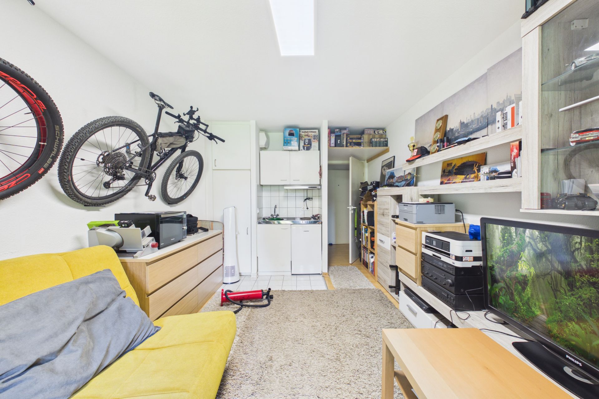Wohnzimmer mit gelbem Sofa, Wandregalen, Fernseher, aufgehängtem Fahrrad und kleiner Küchenzeile.