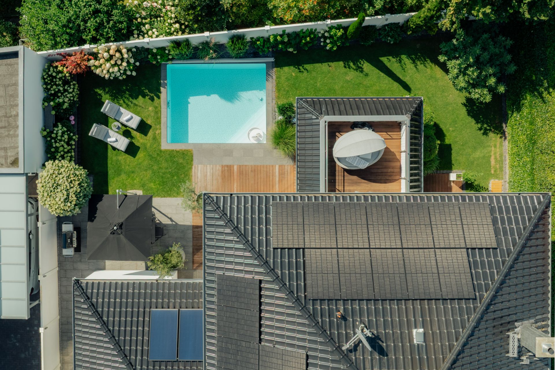 Luftaufnahme eines Gartens mit Pool, Terrasse mit Sonnenschirm und Liegestühlen auf dem Rasen.