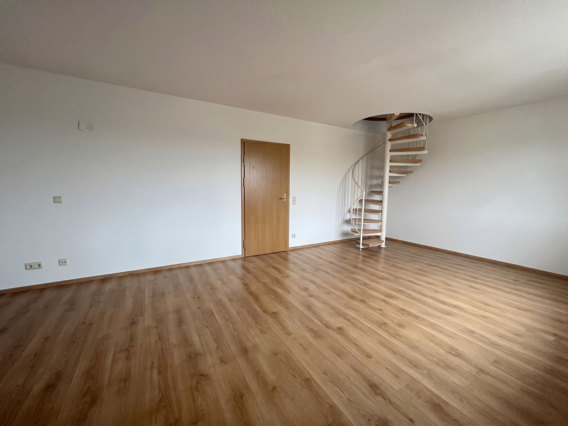 Leerer Raum mit Holzfußboden, weißer Wand, Holztür und Wendeltreppe.