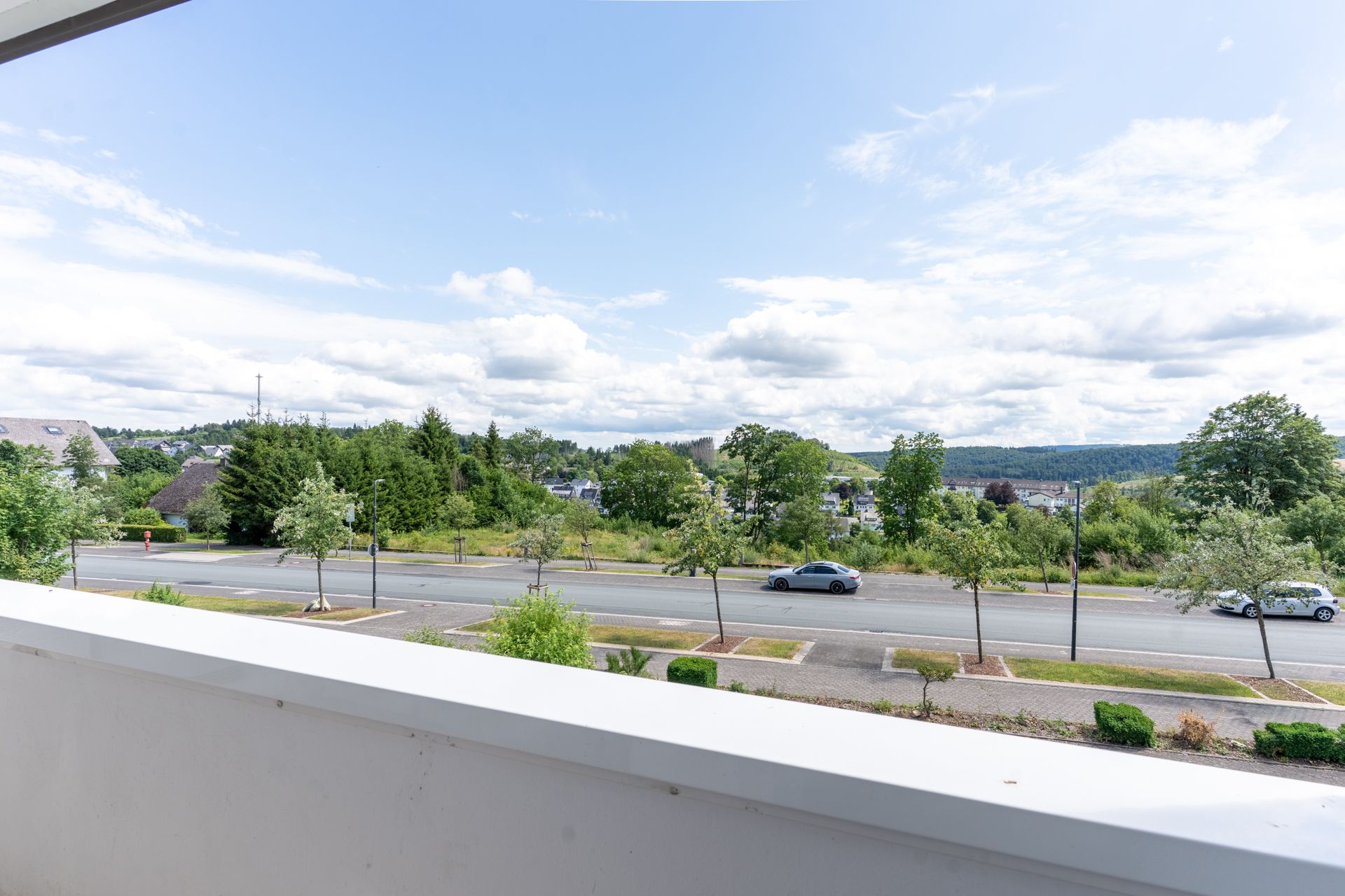 Blick von einem Balkon auf eine Straße mit Autos, Bäumen und grünem Hügel im Hintergrund.