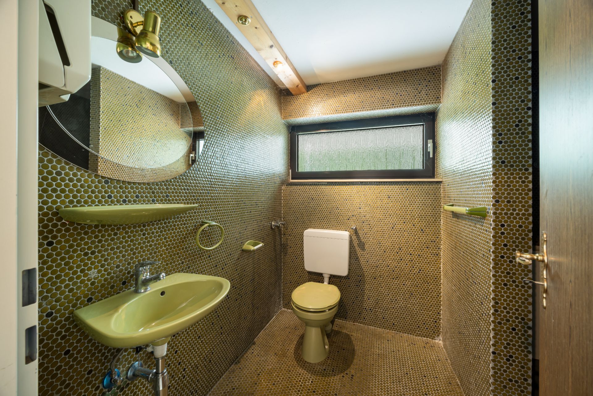 Kleines Badezimmer mit grünem Waschbecken, Toilette und rundem Spiegel, gelben Fliesen an Wand und Boden.