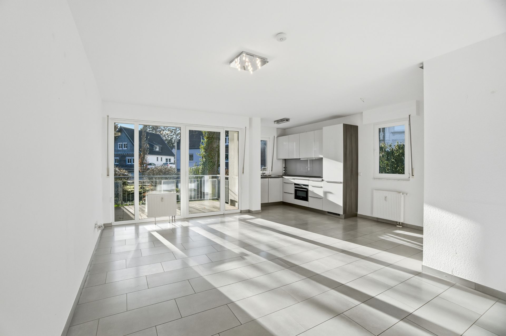 Helle, leere Wohnung mit Fliesenboden, offener Küche und großen Fenstern zur Terrasse.