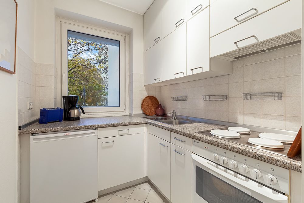 Helle Küche mit weißen Schränken, Herd, Spüle, Kaffeemaschine und Fenster mit Blick nach draußen.