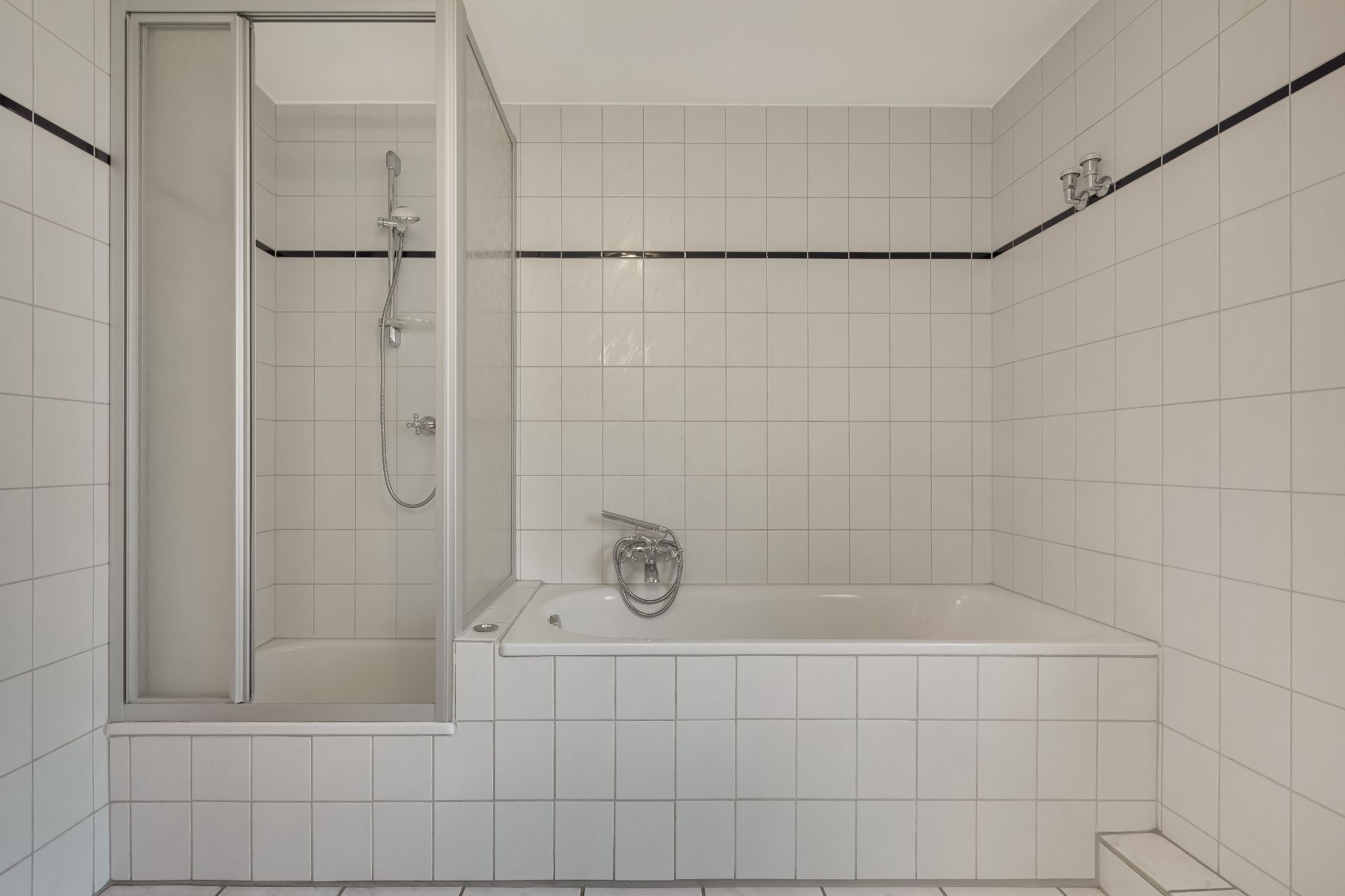 Helles Badezimmer mit Badewanne, Dusche und weißen Fliesen an Wand und Boden.