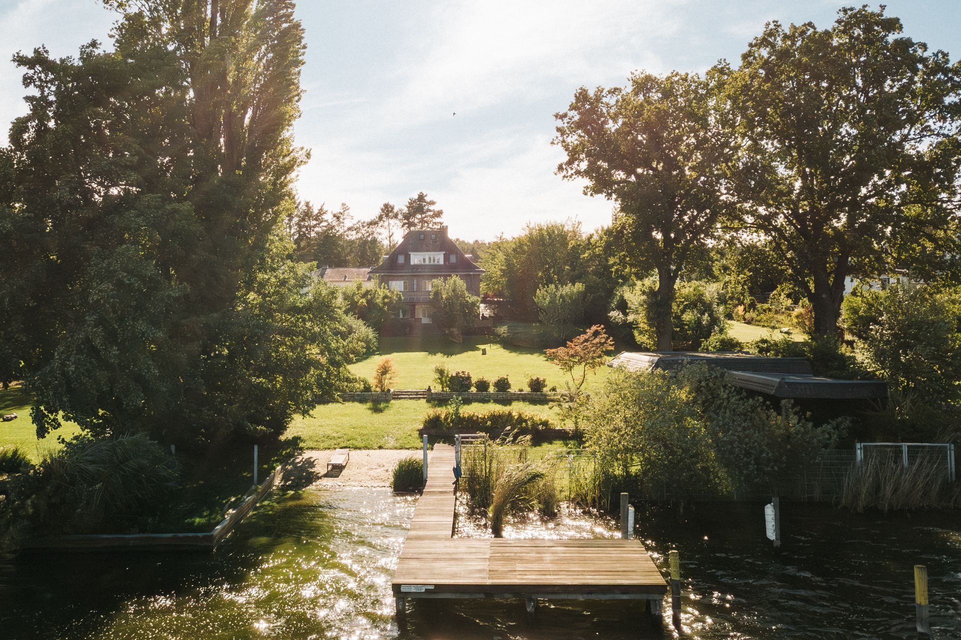 Ein Steg führt zu einem Haus mit großem Garten und Bäumen am Wasser.