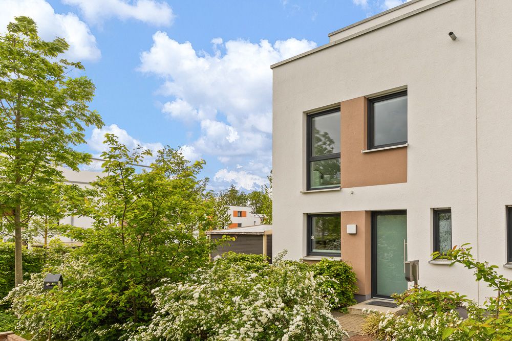 Moderne weiße Hausfassade mit großen Fenstern, umgeben von grünen Büschen und Bäumen.