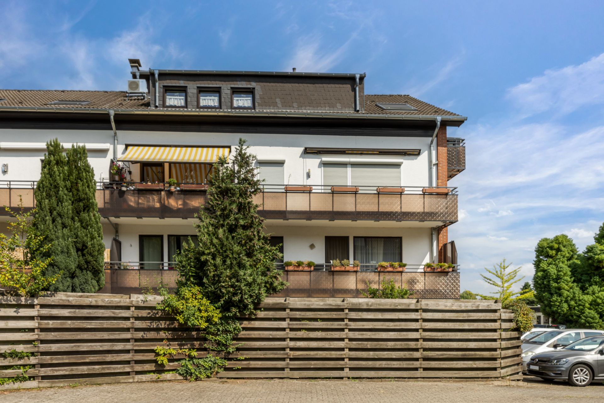 Mehrfamilienhaus mit Balkon, Holz-Zaun und parkenden Autos, umgeben von Bäumen.