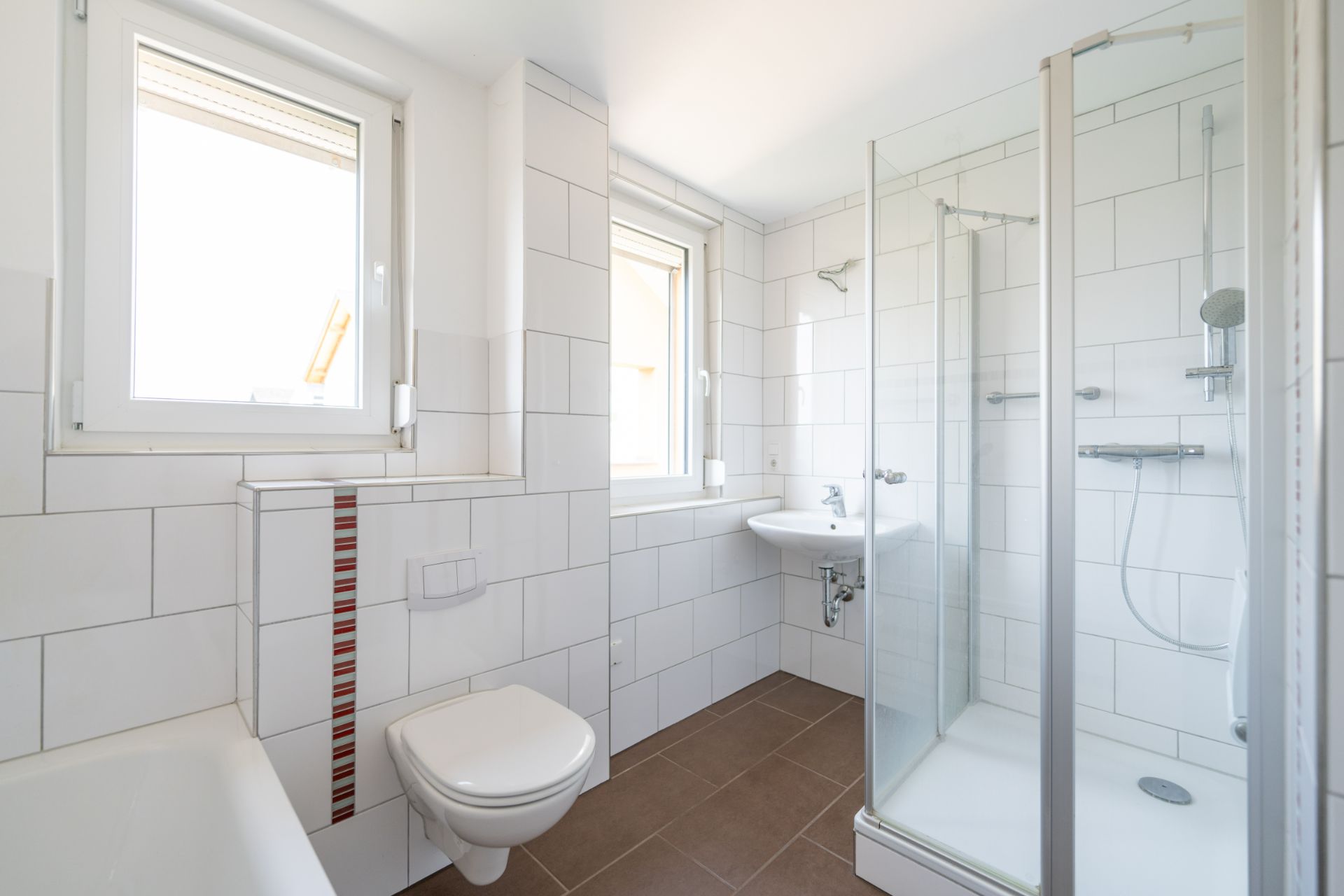 Helles Badezimmer mit Dusche, WC, Waschbecken und zwei Fenstern.
