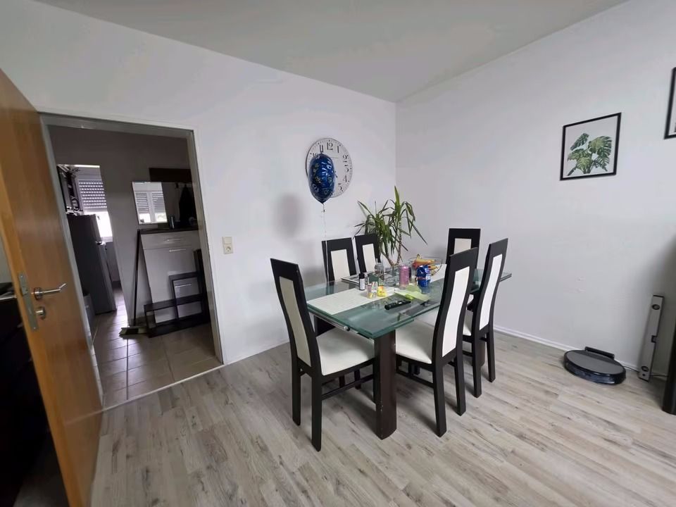Esszimmer mit Glastisch, sechs Stühlen, Uhr, Pflanzen und Dekoration an der weißen Wand.