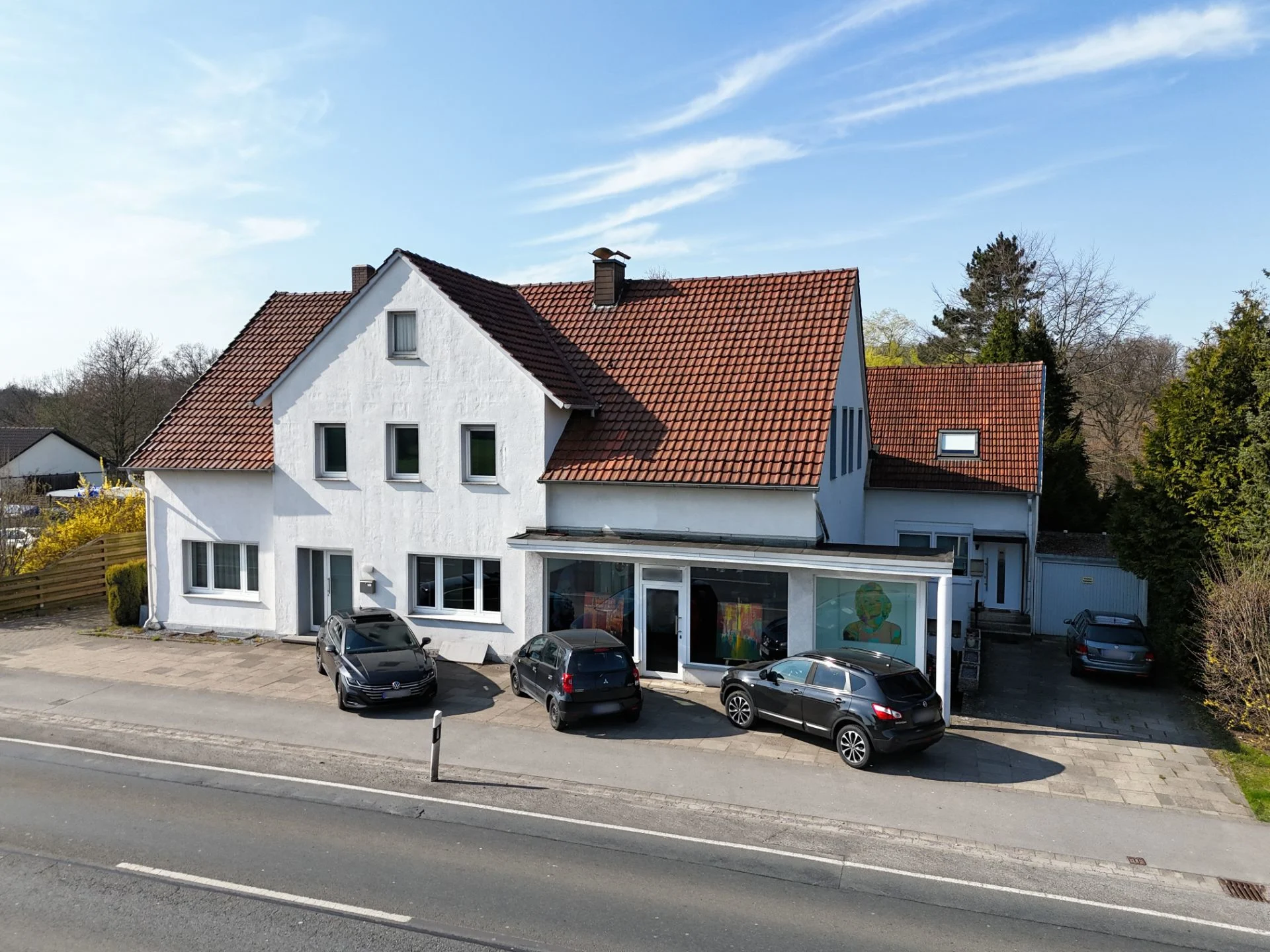 Weißes Wohnhaus mit rotem Dach und mehreren geparkten Autos vor dem Haus an einer Straße.