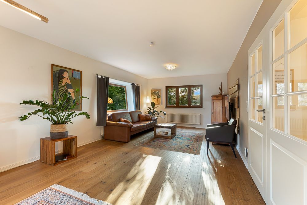Helles Wohnzimmer mit Holzboden, branem Sofa, Sessel, Pflanzen und großem Fenster.