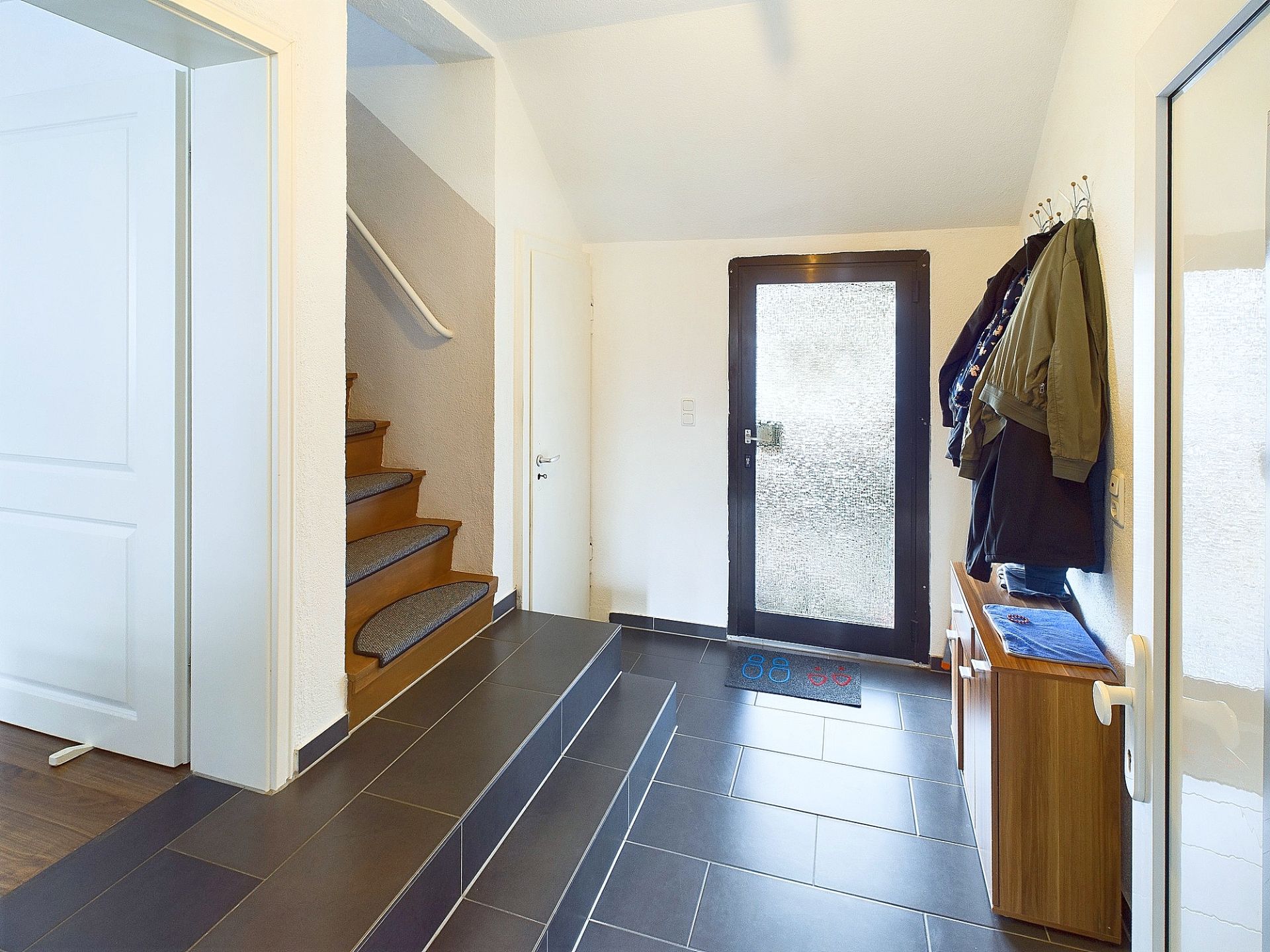 Heller Flur mit Eingangstür, Treppe und Garderobe mit Jacken an der Wand.
