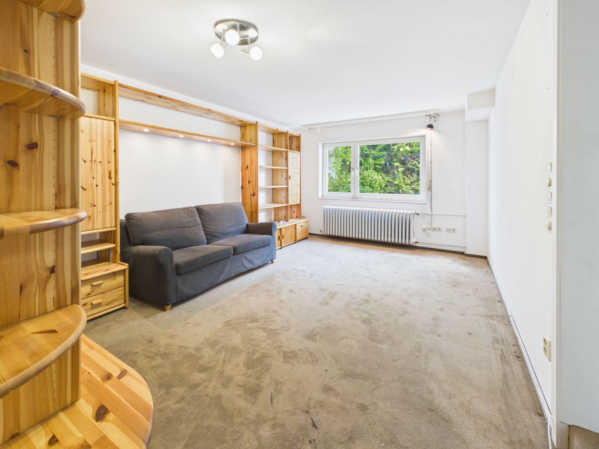 Wohnzimmer mit grauem Sofa, Holzregalen, Teppichboden und großem Fenster mit Blick ins Grüne.