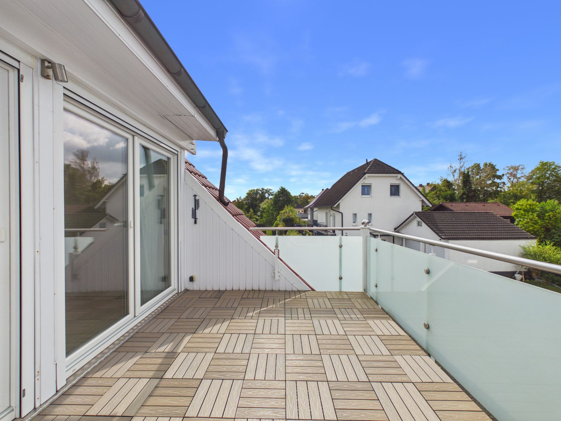 Großer Balkon mit Holzfliesen und Glasgeländer, Blick auf Nachbarhäuser und Bäume.